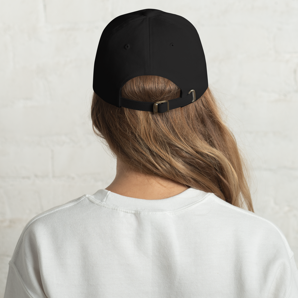 Rear view of a person with long wavy brown hair wearing a plain black baseball cap with a brass buckle closure and a white crewneck sweatshirt against a subtle white brick wall