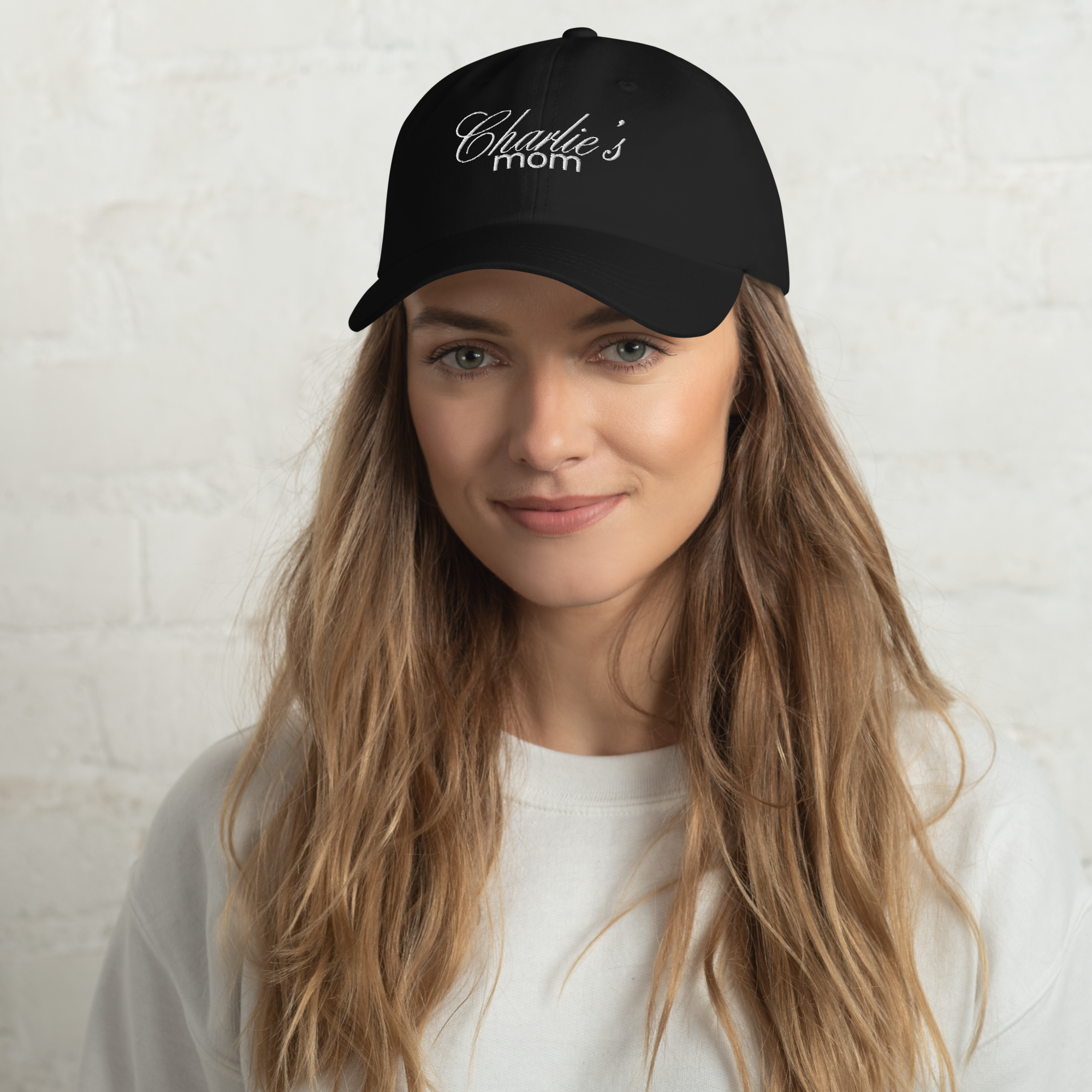 Photograph of a smiling young woman wearing a black baseball cap with "Charlie's mom" embroidered in white script, featuring her long light brown hair and a white sweatshirt, against a bright white brick wall