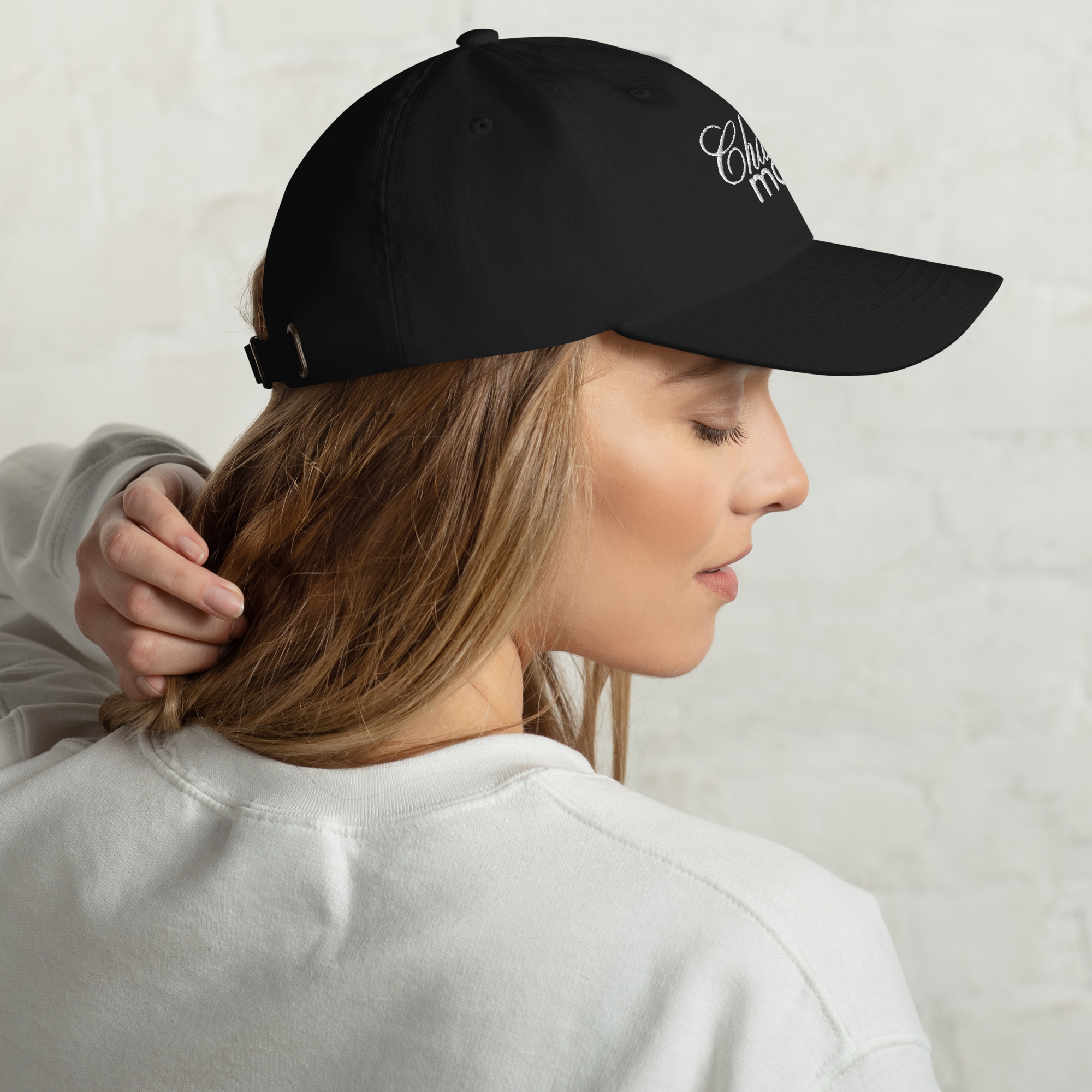 Photograph of a young woman in profile with blonde hair looking away wearing a black baseball cap embroidered with white script 'Chula ma' and a white sweatshirt her hand resting on the back of her head against a white brick wall